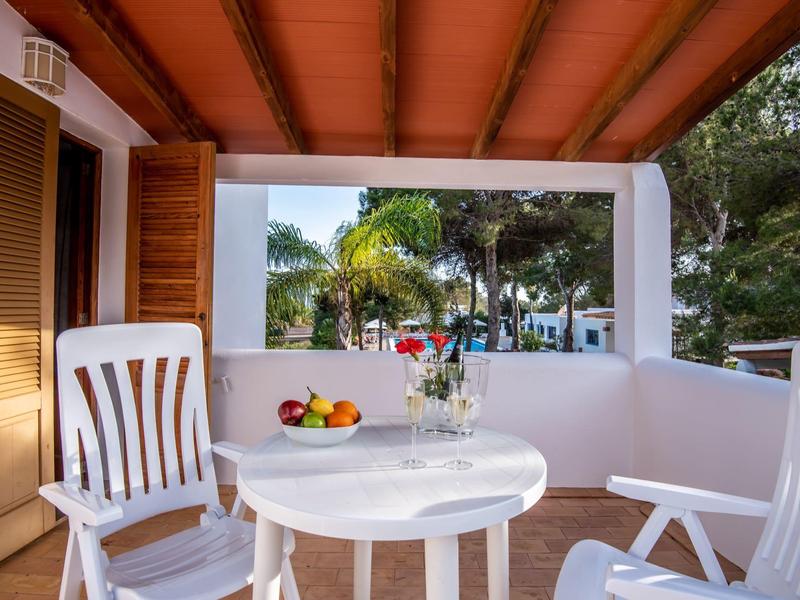 Balcón con mesa y sillas blancas, frutero y botellas de vino, rodeado de palmeras.