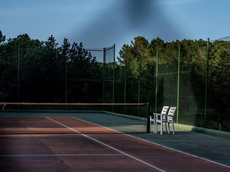 Pista de tenis al aire libre vacía con dos sillas blancas junto a la red y árboles al fondo.