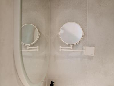 Moderner Badezimmerspiegel mit rundem Vergrößerungsspiegel und weißer Ablage an grauer Wand.