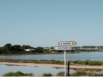 Wegweiser zu La Savina und Es Pujols vor Küstenlandschaft mit Wasser und Vegetation.