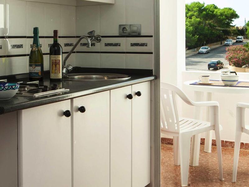 Cocina con armarios blancos y botellas de vino, junto a terraza con mesa y sillas blancas