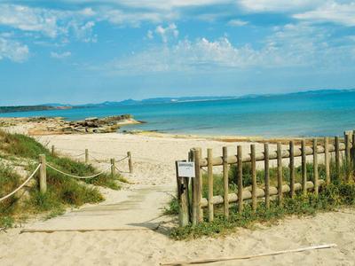 Piaszczysta ścieżka plażowa z drewnianym ogrodzeniem i widokiem na niebieskie morze pod pochmurnym niebem.