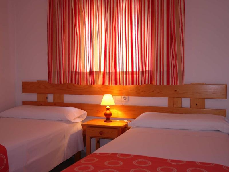 Zwei Einzelbetten mit roten Decken, Nachttisch dazwischen und brennende Lampe vor roten Vorhängen.