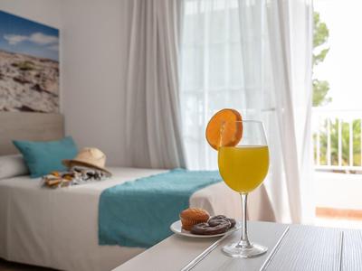 Ein helles Hotelzimmer mit Bett, türkisfarbener Decke und Orangensaft mit Orangenscheibe am Tisch.