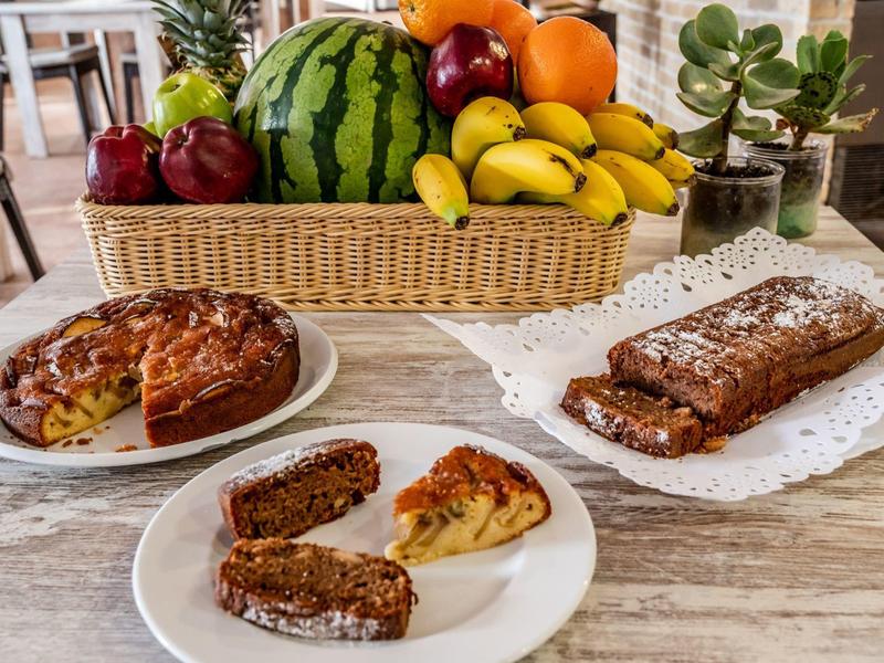 Verschiedene Kuchenstücke und ein Obstkorb mit Melone, Bananen und Äpfeln auf einem Holztisch.