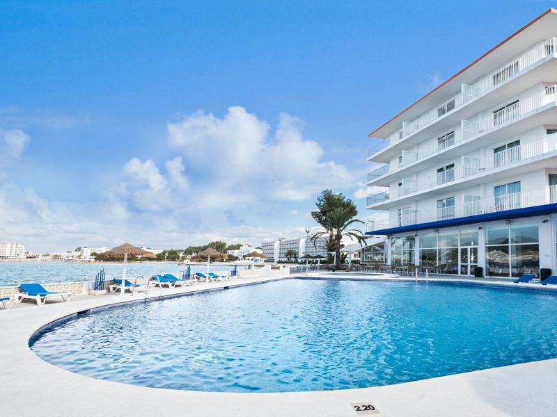 Moderní hotelový bazén s lehátky a výhledem na moře pod modrou oblohou.