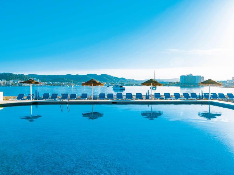 Čistý bazén s lehátky a slunečníky před pobřežní krajinou za jasné oblohy.