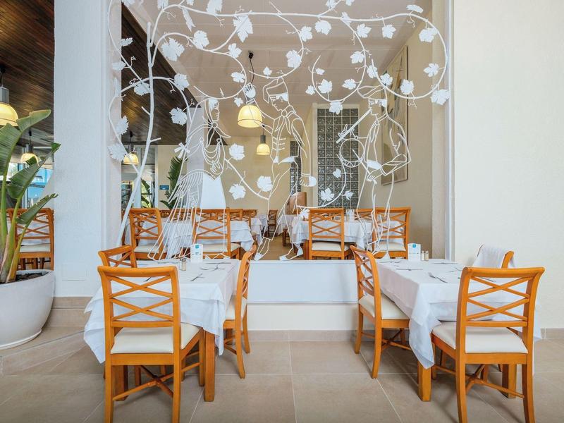 Pohodlná jídelna s dřevěnými stoly, židlemi a dekorativním nástěnným vzorem v světlém prostoru.