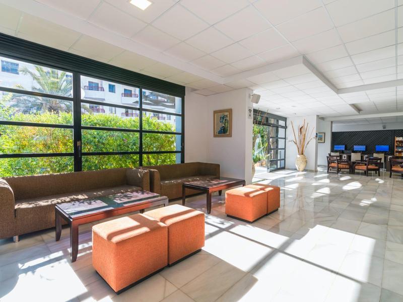 Světlá hotelová lobby s hnědými pohovkami, oranžovými sedátky a velkými okny s výhledem na zeleň.