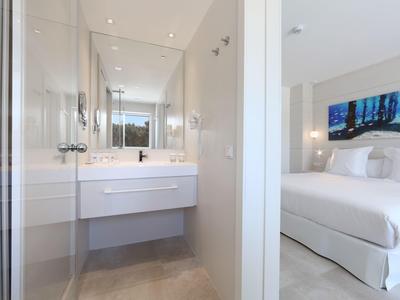 Lichte, moderne hotelkamer met wit bed en aangrenzende badkamer met wastafel en spiegels.