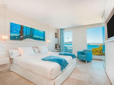 Hell eingerichtetes Schlafzimmer mit Doppelbett, blauem Sessel und Meerblick durch große Fenster.