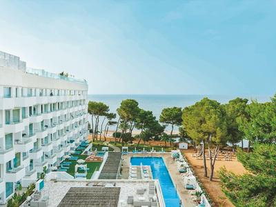 Vista di un hotel con piscina, circondato da alberi e mare sullo sfondo sotto un cielo limpido.