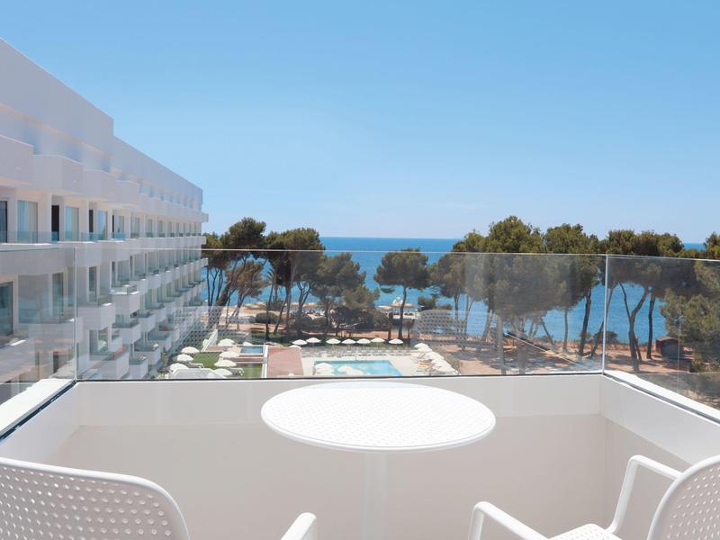 Balkon met tafel en stoelen met uitzicht op zwembad, bomen en zee bij heldere lucht.