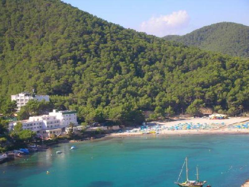 Beach with clear water, surrounded by forested hills and white buildings along the shore.