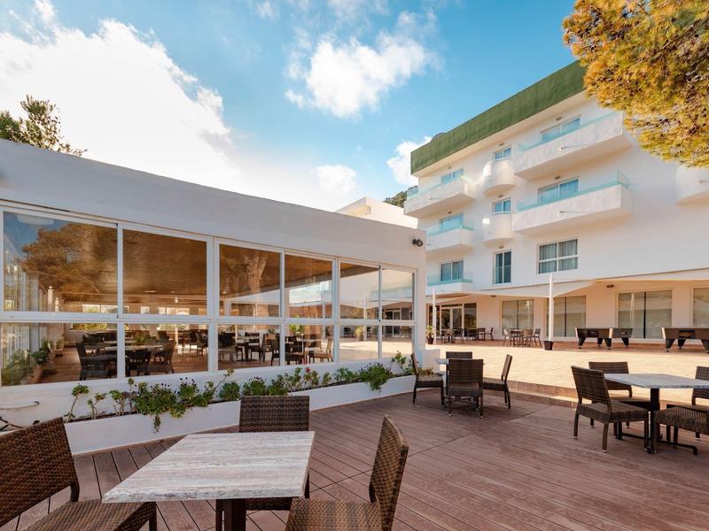 Hotel terrace with wooden seating areas in front of a multi-story white building under sunlight