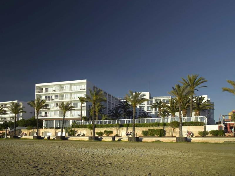 Moderní bílý hotel s palmami na písčité pláži při západu slunce.