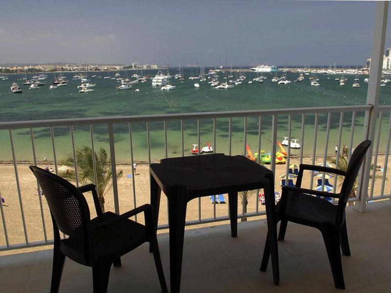 Balkon se stolem a židlemi s výhledem na pláž a moře s loděmi.