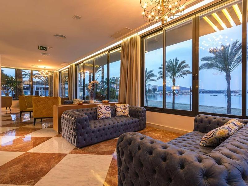 Elegantní hotelový salon s pohovkami a výhledem na moře a palmy při západu slunce.