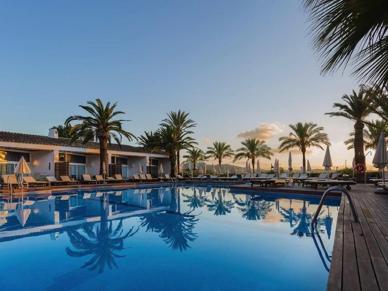 Hotelový bazén s palmami pod modrou oblohou při západu slunce.