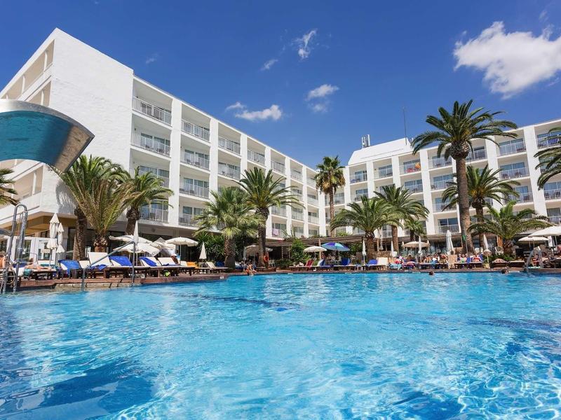 Venkovní bazén moderního hotelu obklopený palmami pod jasně modrou oblohou.