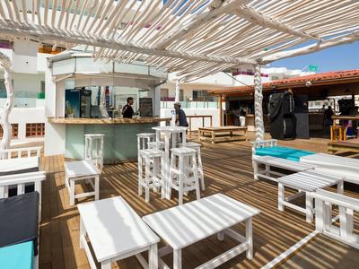 Freiluftbar mit weißen Holzmöbeln, Holzboden und geflochtenem Dach, helle und offene Atmosphäre.