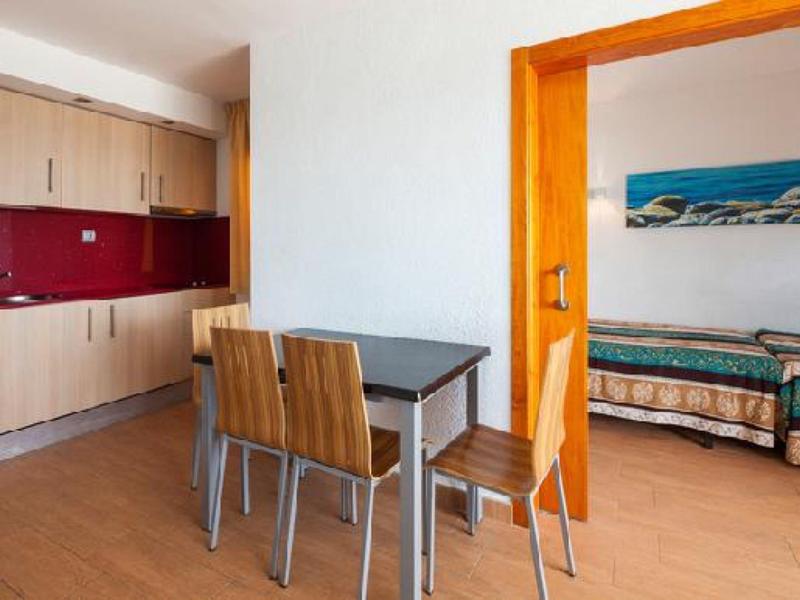 Moderner Raum mit kleinem Essplatz, Holztisch, vier Stühlen, angrenzender Küche und Schlafzimmer mit Betten.