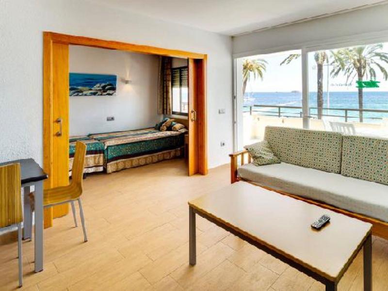 Helles Wohnzimmer mit Sofa, Tisch, Stühlen, Schlafzimmer und Meerblick durch große Fenster.