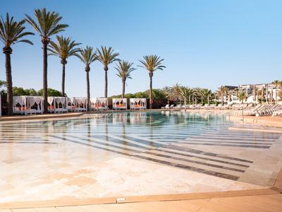 Large hotel pool with lounge chairs, palm trees, and clear blue sky.