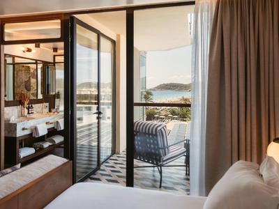 Hotel room with open door to terrace with chair and sea view in sunlight.