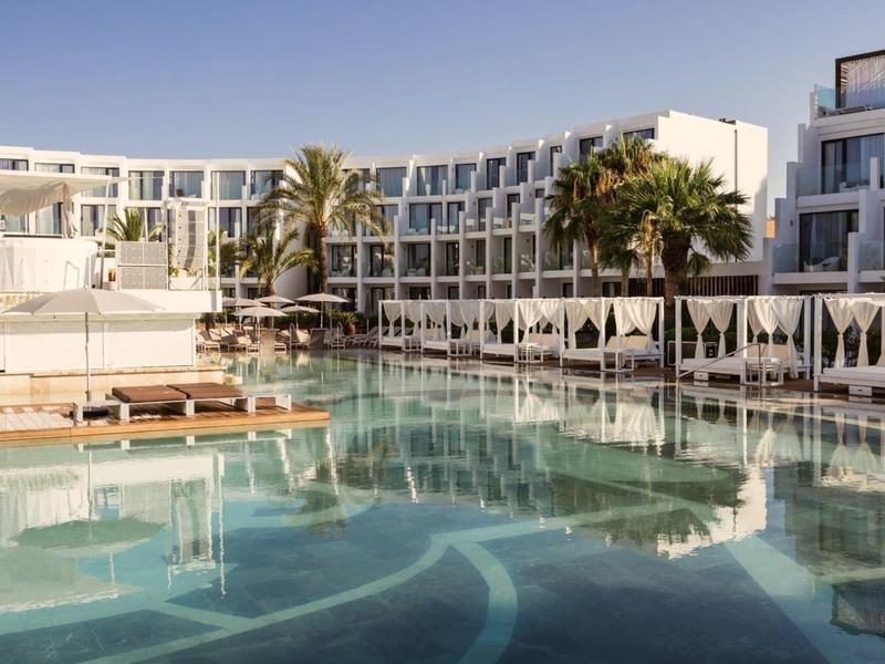 Modern hotel complex with large pool, loungers, and white umbrellas under clear sky.