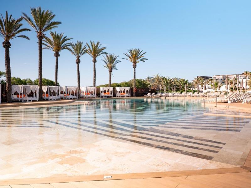 Large hotel pool with lounge chairs, palm trees, and clear blue sky.
