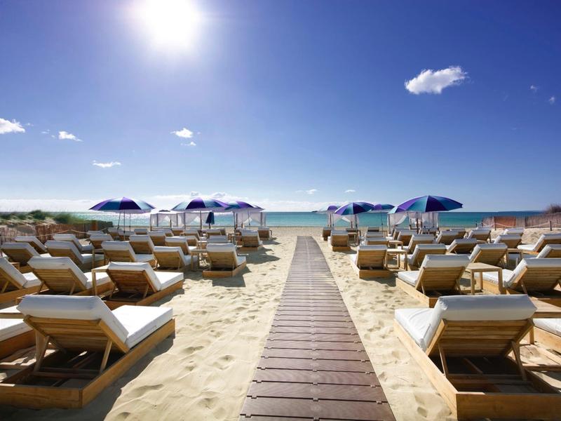 A wooden path leads to a sunny beach with lounge chairs and blue umbrellas by the sea.