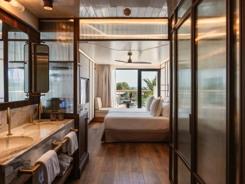 Modern hotel room with wooden floor, double bed, and bathroom featuring two sinks and large mirrors.