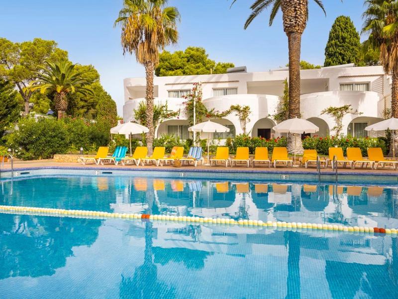 Klare Poolanlage mit Liegestühlen, Sonnenschirmen und weißen Gebäuden im Hintergrund bei Sonnenschein.