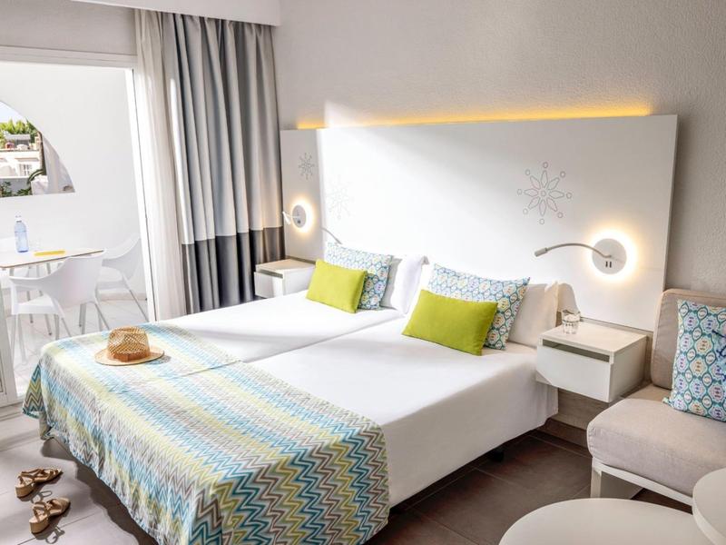 Helles Schlafzimmer mit Doppelbett, buntem Bettüberwurf, grünen Kissen und Balkon mit Sitzbereich.