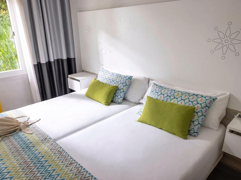 Zwei weiße Einzelbetten mit bunten Kissen und Decken in einem hellen, modernen Schlafzimmer.