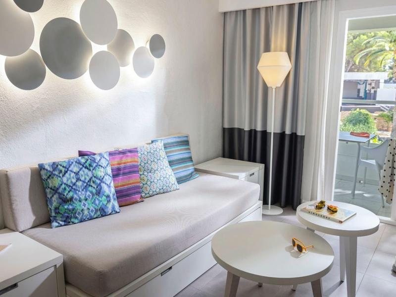 Gemütliches Wohnzimmer mit hellgraßer Couch, bunten Kissen, runden Tischen und Zugang zum Balkon.