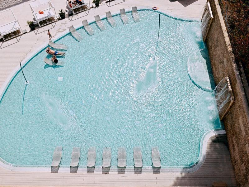 Piscina con tumbonas, chorros de agua y pared con efecto de agua en área del hotel.