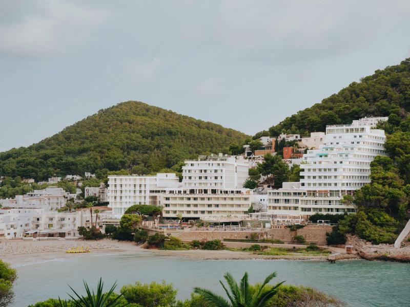 Edificios de hotel blancos en una colina con vegetación verde y playa en primer plano.