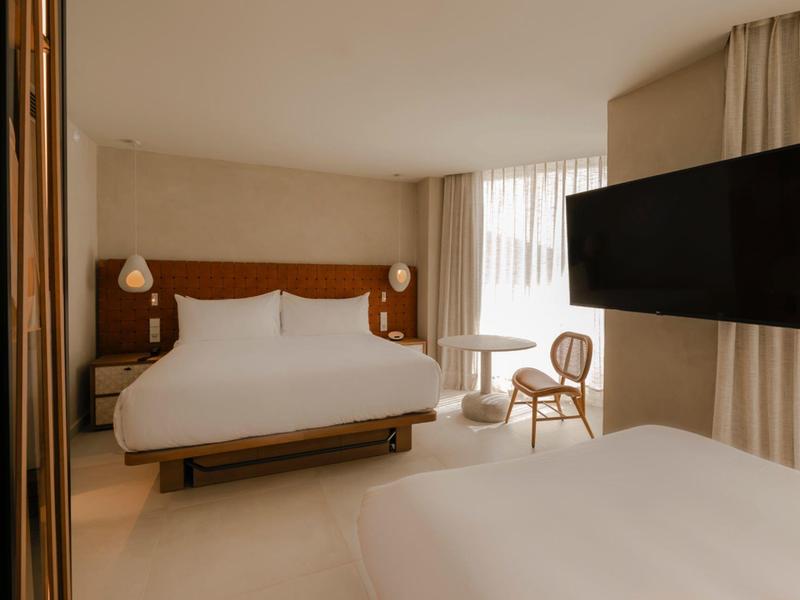 Habitación de hotel luminosa con dos camas, silla, mesa, ventana grande y televisor en la pared.