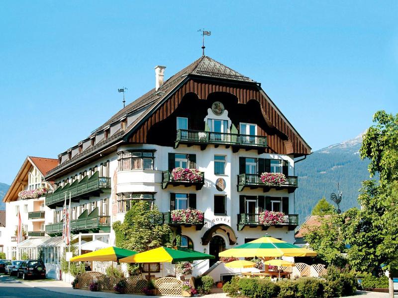 Traditional Alpine hotel with balconies and colorful awnings, surrounded by mountains and green foliage.