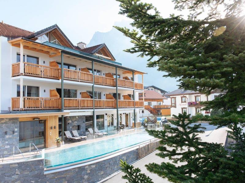 Modern hotel with wooden balconies and outdoor pool, surrounded by mountains and trees.