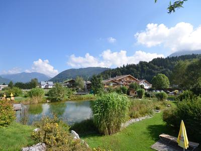Grüner Garten mit Teich, Büschen und Häusern vor bewaldeten Bergen bei klarem Himmel