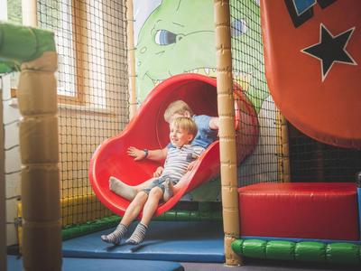 Dos niños bajan por un tobogán rojo en un área de juegos interior colorida.