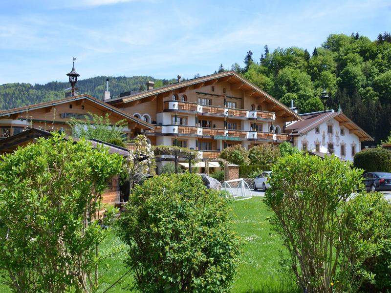 Hotel de estilo alpino con arbustos verdes en primer plano y colinas boscosas al fondo.