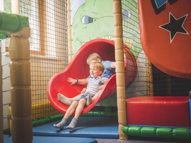 Dos niños bajan por un tobogán rojo en un área de juegos interior colorida.
