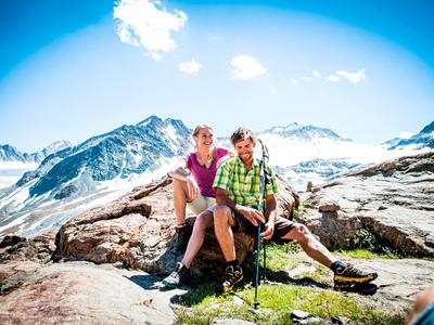 Ein Paar sitzt auf Felsen vor einer schneebedeckten Berglandschaft bei klarem Himmel.