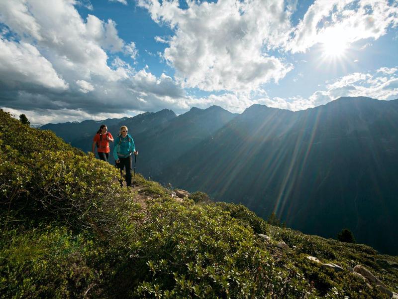 Dos excursionistas están en un prado verde en la montaña mirando un valle iluminado por el sol y cielo nublado.