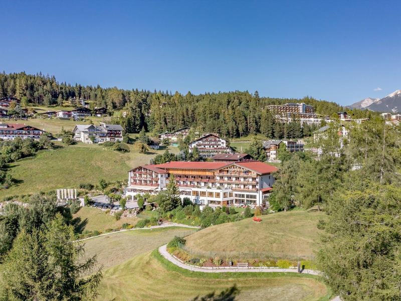 Grünes Tal mit mehreren Gebäuden, bewaldeten Hügeln und Bergen im Hintergrund unter blauem Himmel.