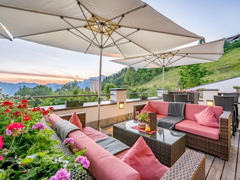 Terrassenmöbel aus Rattan mit roten Kissen unter großem Sonnenschirm, Blumen und Berge im Hintergrund.